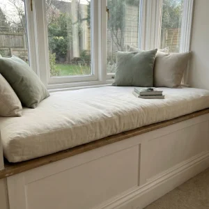 Custom-fitted cream linen cushion on a white wooden bay window seat, styled with sage green and beige throw pillows.